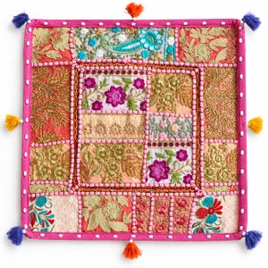 Puede incluir: Una funda de cojín decorativa cuadrada con un diseño de patchwork de patrones florales bordados en rosa, dorado, turquesa y naranja. El borde es rosa brillante con detalles de conchas blancas y borlas azules y naranjas en las esquinas.