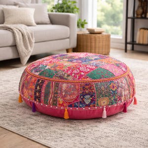 Puede incluir: Un puf de suelo redondo y colorido con un diseño de patchwork. El puf presenta una vibrante mezcla de telas rosas, verdes, naranjas y moradas. Tiene una base rosa con borlas decorativas, añadiendo un toque bohemio a la decoración del hogar.