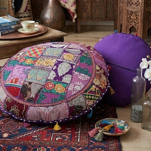 May include: Two round floor cushions. One is a patchwork of floral and geometric patterns in purple, pink, and gold. The other is solid purple. Tassels adorn the edges. The cushions rest on a patterned rug.