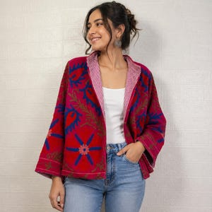 Op de afbeelding: Rode jas met blauwe en groene bloemenborduursels. De jas heeft een gestreepte voering en wordt gedragen over een witte top en een blauwe jeans. Het ontwerp van de jas heeft een centraal bloemmotief.