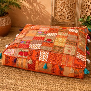 May include: A square floor cushion featuring a patchwork design of various orange, gold, and red fabric panels with intricate embroidery, floral patterns, and metallic thread accents. The edges are decorated with small, colourful hanging tassels.