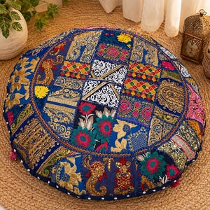 May include: A round floor cushion featuring a patchwork design of blue fabric panels. The surface is decorated with intricate gold embroidery, floral patterns, sequins, and small mirrors. The edges are finished with a white pom-pom trim detail.
