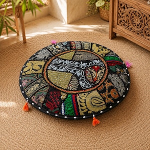 May include: A round floor cushion featuring a patchwork design of various fabrics in black, gold, green, and red. The surface is decorated with intricate embroidery, metallic thread, and small mirrors. The edges are finished with small orange tassels.