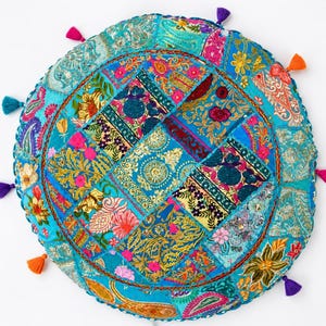 Könnte beinhalten: Ein runder Patchwork-Bodenkissen in Türkis, Rosa, Gold und Lila. Das Kissen hat ein zentrales Design aus quadratischen Flicken mit Blumen- und Paisley-Mustern. Bunte Quasten säumen den Rand.