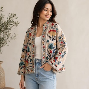 Può includere: Giacca beige ricamata con motivi floreali colorati. La giacca ha un collo alto e maniche lunghe. Il ricamo comprende fiori e foglie rosse, blu, gialle e verdi. La giacca è indossata sopra un top bianco e jeans blu.