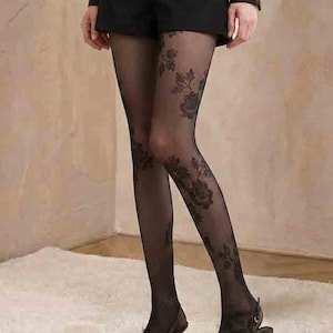Pode incluir: Meias-calças pretas transparentes com padrão floral, usadas com calções pretos e sapatos de salto. As meias-calças apresentam um design floral escuro detalhado. Os sapatos são pretos com bico fino.