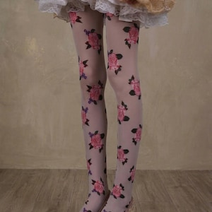 Puede incluir: Medias rosas claras adornadas con un patrón repetido de rosas rosas y hojas verdes. Las medias se usan con tacones rosas a juego con lazos. El atuendo se completa con una falda blanca con ribete de encaje y un top rosa y beige.