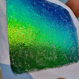 May include: A piece of white paper displaying a colour gradient, shifting from blue to green and yellow. The surface has a glittery, textured look, with the colours blending. The paper is held by a finger.