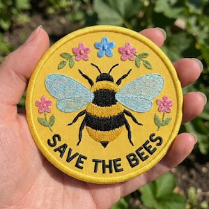 May include: A circular yellow embroidered patch featuring a central bumblebee with black and yellow stripes and light blue wings. The design includes small pink and blue flowers and the black embroidered text SAVE THE BEES along the bottom edge.