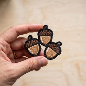 May include: An embroidered patch featuring three brown acorns with dark brown caps and tan bodies, each outlined in black thread. The design includes small white highlights on the side of each acorn nut to provide a sense of depth and texture.