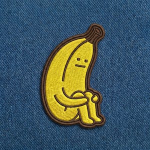 May include: Embroidered patch featuring a yellow banana with a sad face, hugging its knees. The banana has a brown outline and stem, set against a blue denim background. The patch is a fun, quirky accessory.
