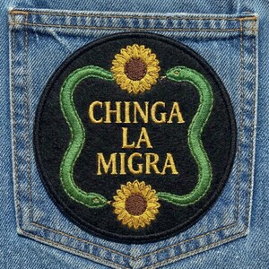 May include: A round black patch with the text "CHINGA LA MIGRA" in gold lettering. The patch features a green snake design and two yellow sunflowers. The patch is sewn onto the back pocket of a pair of blue jeans.