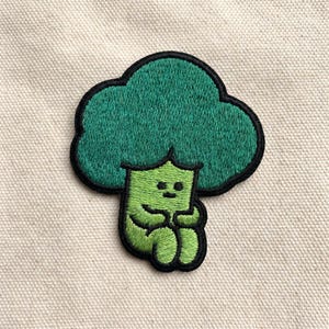 May include: Embroidered patch featuring a cartoon broccoli character with a smiling face and arms crossed. The broccoli is green with a black outline, and the patch is on a beige fabric background. The patch is designed to be ironed on or sewn onto clothing or accessories.