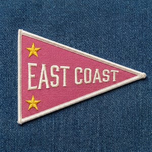 May include: A triangular pink embroidered patch with a white border. The text EAST COAST is stitched in white capital letters across the center. Two yellow five-pointed stars are positioned above and below the text on the left side.
