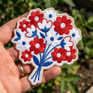 May include: An embroidered patch featuring a bouquet of flowers in red, white, and blue thread. The design includes several red and white blossoms with blue centers, accented by blue stems and leaves, all bordered by a scalloped cream-colored edge.