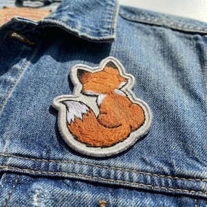 May include: Embroidered patch featuring a stylized orange fox with a white-tipped tail, set against a denim jacket. The fox is outlined in gray, adding a touch of detail to the design. This patch is ideal for adding a personalized touch to clothing or accessories.