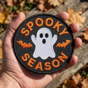 May include: A circular embroidered patch with a black background featuring a white ghost in the center, two orange bats on the sides, and the orange text SPOOKY SEASON arched around the ghost. The patch has a black stitched border edge.