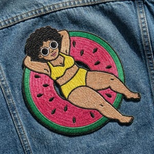 May include: Embroidered patch featuring a woman in a yellow bikini and sunglasses, relaxing on a watermelon-shaped float. The patch has a pink and green border, with black seeds on the watermelon. The patch is attached to a denim jacket.