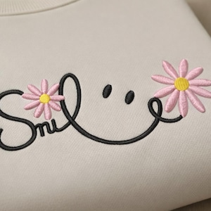 May include: Cream-colored sweatshirt with embroidered design. The design features the word "Smile" in black script, with two pink daisies with yellow centers. The "i" in smile is replaced with two black dots.