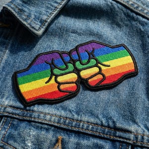 May include: A colorful embroidered patch featuring two fists bumping, with each fist displaying a rainbow gradient. The patch is outlined in black and is set against a blue denim jacket.