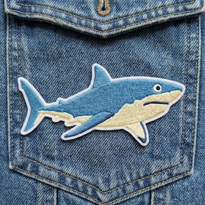 May include: A blue and white embroidered shark patch is sewn onto a denim jacket pocket. The shark has a light blue body, white accents, and a beige belly. The denim is a medium wash.