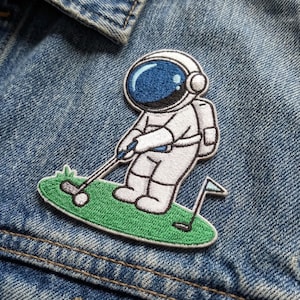 May include: Embroidered patch featuring an astronaut playing golf. The astronaut is wearing a white spacesuit with a blue visor, swinging a golf club on a green patch of grass. A small flag marks the hole. The patch is on a denim jacket.