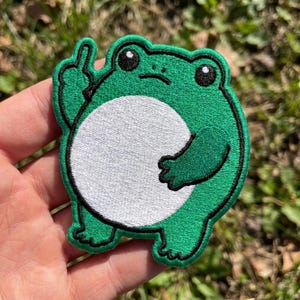 May include: An embroidered patch shaped like a round green frog with a large white belly, black eyes, and a small mouth. The frog is depicted with one arm raised, extending its middle finger upward, outlined in thick black thread throughout.