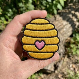 May include: An embroidered patch shaped like a yellow beehive with horizontal black stitching lines and a small pink heart centered on the front. The patch features a thick black border outlining the rounded, tiered structure of the hive.