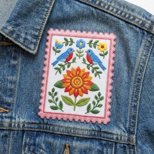 May include: Embroidered patch featuring a vibrant design of two bluebirds perched on branches above a sunflower. The patch has a pink scalloped border and is set against a denim jacket. The patch includes colorful flowers and green leaves.