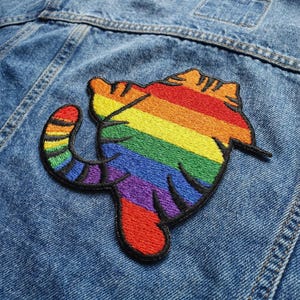 May include: Embroidered patch in the shape of a cat, featuring a rainbow design. The patch is sewn onto a blue denim jacket. The cat's body is composed of horizontal stripes in the colors of the rainbow.