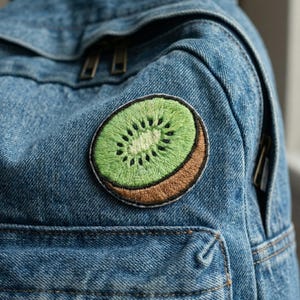May include: A denim jacket with a round kiwi fruit patch. The patch features a green center with black seeds and a brown outer edge, stitched onto the blue denim fabric. The jacket has a pocket and zipper detail.