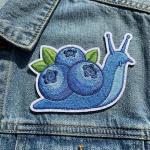May include: Embroidered patch featuring a blue snail with a shell made of three blueberries and green leaves. The patch has a white border and is set against a denim jacket.