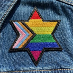 May include: Embroidered patch in the shape of a six-pointed star, featuring a rainbow design with black border. The patch includes the colors of the Pride flag and the Transgender Pride flag. The patch is sewn onto a denim jacket.
