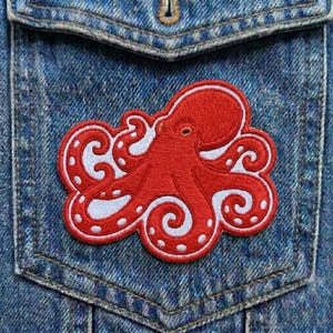May include: A red octopus patch with white accents is sewn onto a denim pocket. The octopus design features detailed tentacles and a white border. The patch is a decorative element for clothing or accessories.