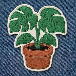 May include: An embroidered patch featuring a potted monstera plant with four distinct green leaves and a brown terracotta-colored pot. The design is outlined with a thick cream-colored border, showcasing detailed stitching throughout the plant and pot.