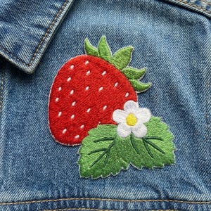 May include: Embroidered patch featuring a vibrant red strawberry with white seeds, green leaves, and a white flower with a yellow center. The patch is sewn onto a blue denim jacket, showcasing a playful, summery design.