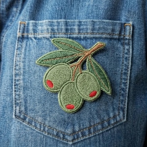 May include: Embroidered olive branch patch with green olives and red accents, attached to a blue denim pocket. The patch features detailed stitching and a naturalistic design, perfect for adding a touch of nature to clothing or accessories.