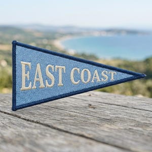 May include: A triangular pennant flag featuring a light blue fabric base with a dark blue stitched border. The text EAST COAST is embroidered in large, cream-colored capital letters across the center of the light blue surface.