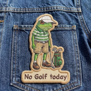 May include: Embroidered patch featuring a green frog in a golf outfit, wearing a white cap and holding a golf club. The patch includes the text "No Golf today" in a tan border. The patch is attached to a denim jacket.