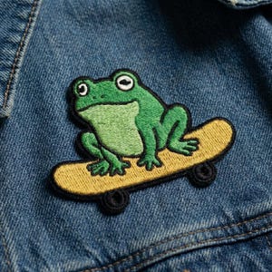 May include: Embroidered patch featuring a green frog riding a yellow skateboard. The frog has white eyes and is positioned on the skateboard with black wheels. The patch is sewn onto a blue denim jacket.