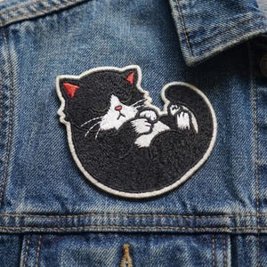 May include: Embroidered patch featuring a black and white cat in a curled-up sleeping position. The cat has red accents on its ears and nose, with white whiskers. The patch is outlined in white and is on a denim jacket.