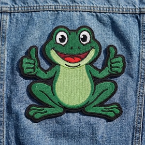 May include: A green frog patch with a smiling face and thumbs up. The frog has a light green belly, dark green outline, and black border. The patch is on a blue denim jacket.