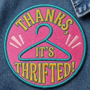 May include: A circular embroidered patch with a pink background and a teal border. The patch features a teal clothes hanger graphic in the center with the yellow embroidered text THANKS, IT'S THRIFTED! and small light pink accent lines.