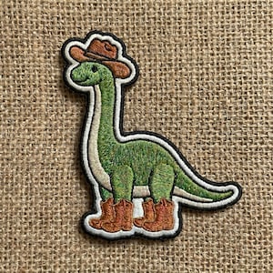 May include: Embroidered patch featuring a green dinosaur wearing a brown cowboy hat and brown cowboy boots. The dinosaur has a long neck and a white belly. The patch has a black border and is on a burlap background.