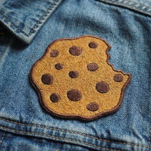 May include: Embroidered cookie patch with brown chocolate chips and a bite taken out, sewn onto a denim jacket. The patch is tan with a brown border and is a fun accessory for clothing.