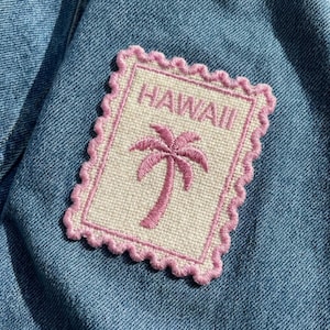 May include: Embroidered patch featuring a pink palm tree and the word "HAWAII" on a cream-colored background. The patch has a pink scalloped border and is set against a blue denim fabric.