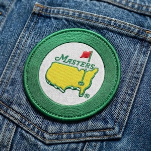 May include: A circular embroidered patch featuring the Masters Tournament logo. The patch has a green border, a white interior, and a yellow map of the United States with a red flag. The word "Masters" is written in cursive.