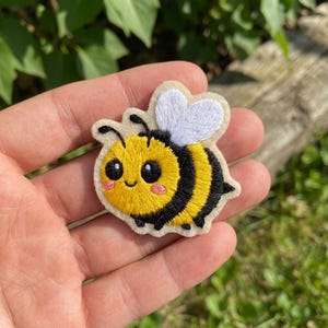 May include: A small embroidered patch featuring a cartoon bee with a yellow and black striped body, large black eyes, pink cheeks, and white heart-shaped wings. The design is stitched onto a cream-colored fabric backing with a scalloped edge.