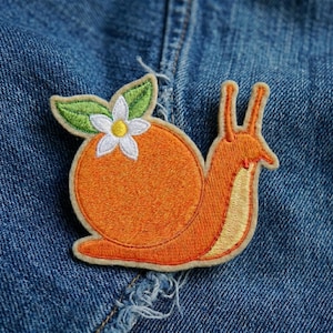 May include: Embroidered patch featuring an orange snail with an orange body, a cream-colored belly, and a flower on its back. The flower has white petals and a yellow center. The patch is on a blue denim background.