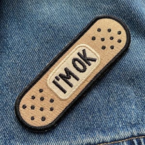 May include: A tan and black embroidered patch in the shape of a bandage. The patch has the words "I'M OK" embroidered in black on a cream-colored rectangle. The patch is on a blue denim background.